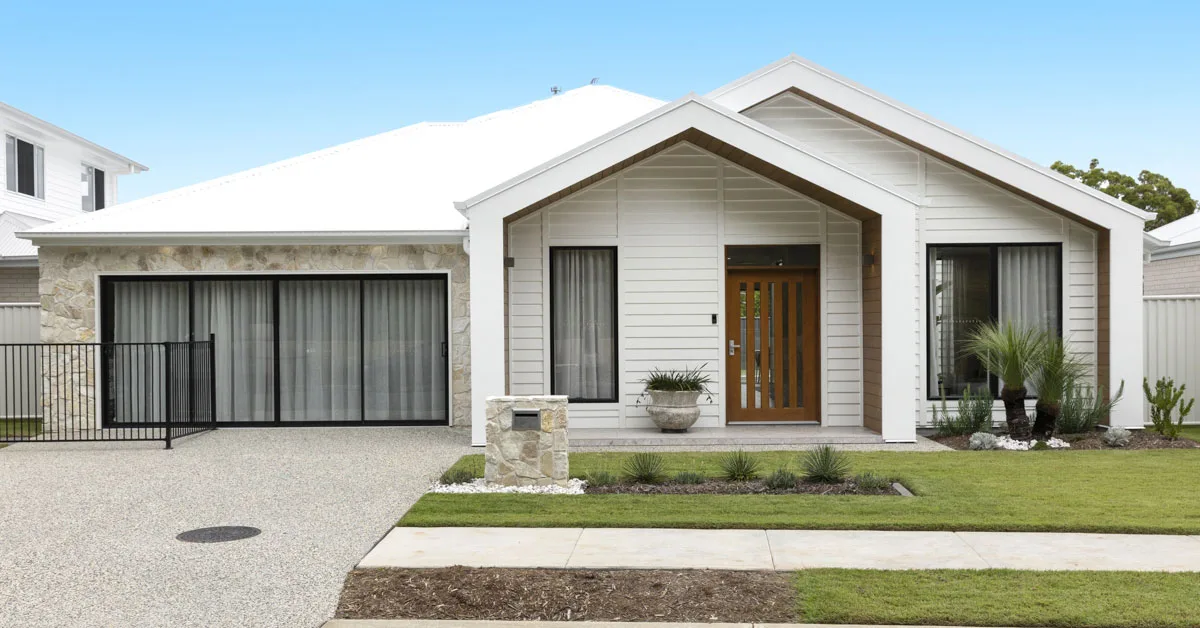 Modern white house with a gabled roof, stone accents, and a wooden front door. Lush green lawn, glass sliding doors, and a clear blue sky.