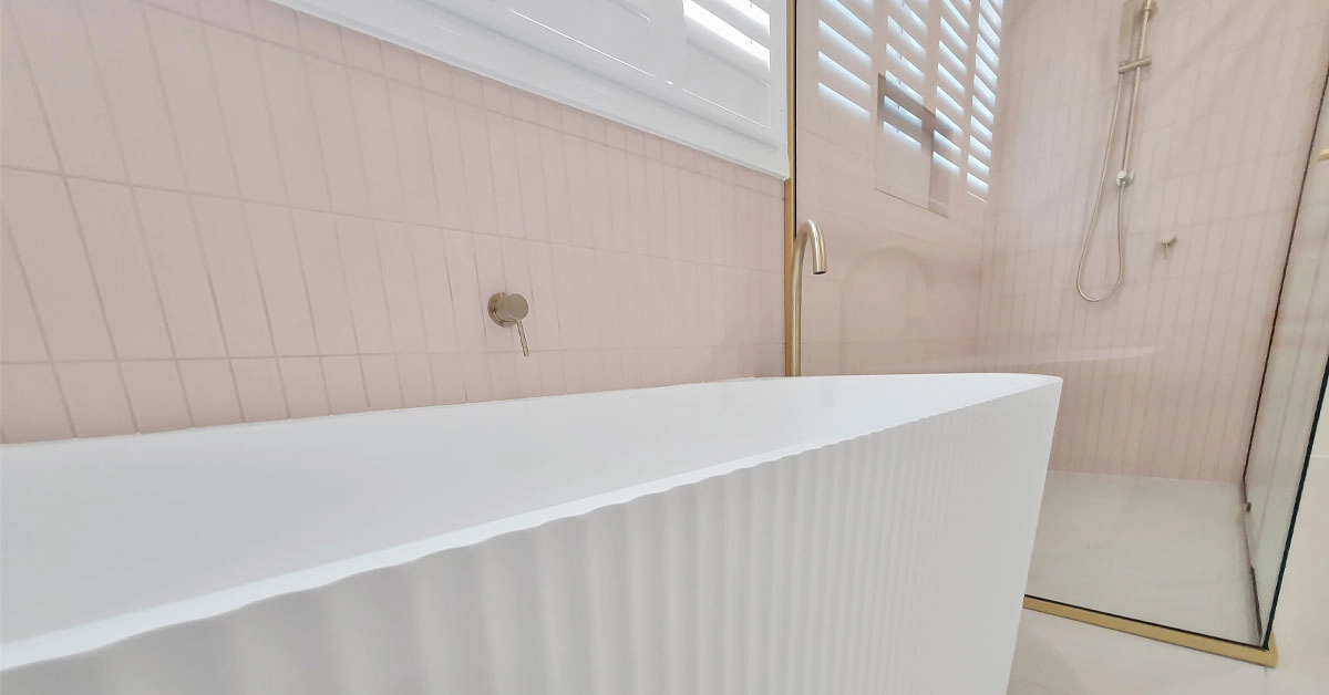 Minimalist bathroom featuring a white fluted bathtub with brass fixtures. Pale pink tiles and frosted glass shower add a serene, elegant touch.