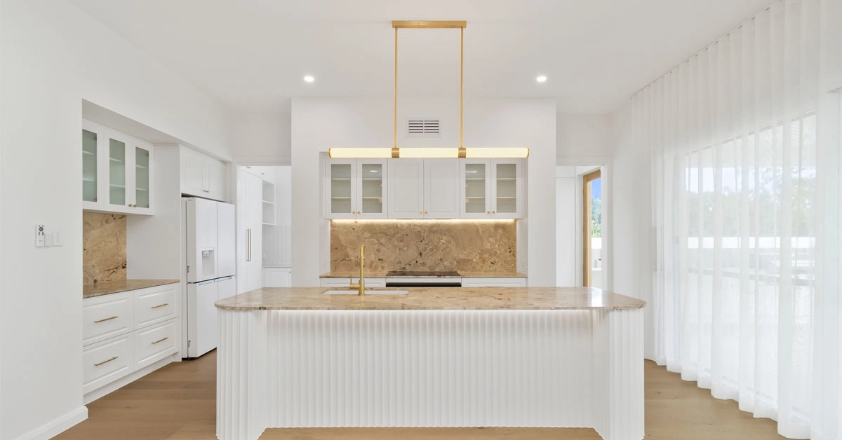 Modern kitchen with white cabinetry, marble countertops, and gold fixtures. Large island with a sleek hanging light fixture. Bright and airy feel.