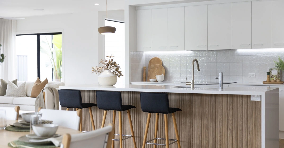 Modern kitchen with wooden floors, white cabinets, and a sleek island featuring black barstools. A vase with leaves adds warmth and elegance.