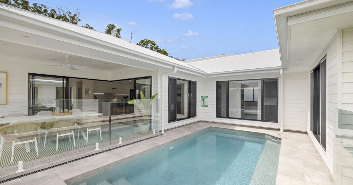 Modern outdoor area featuring a clear blue pool, surrounded by white walls and glass fencing. Dining area with wicker chairs under a covered patio. Bright, serene atmosphere.