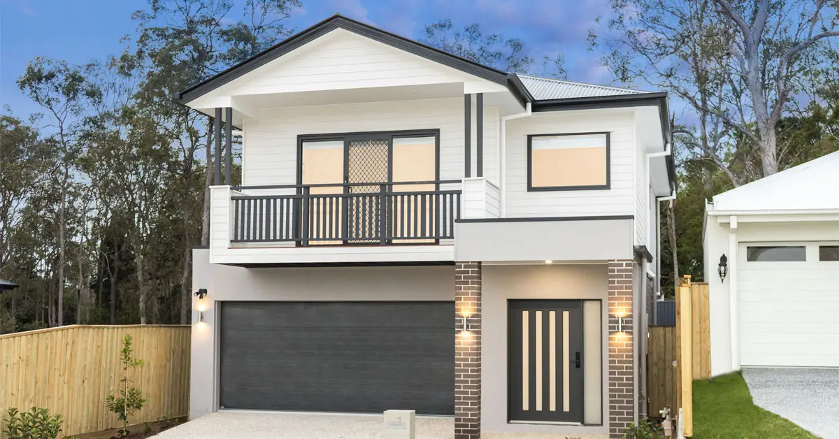 Modern two-story house with a gray garage door, a balcony with black railing, and brick accents. Surrounded by trees and dusk lighting, conveying tranquility.