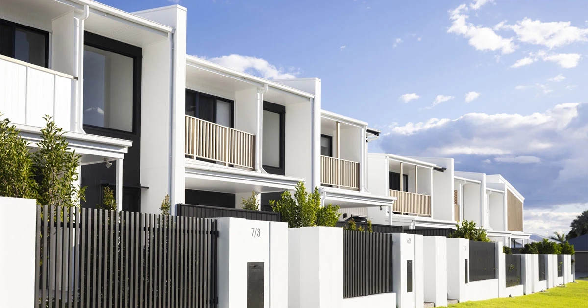 Modern white townhouses with large windows and balconies line a street, bordered by a sleek black fence. The sky is clear with a few clouds.