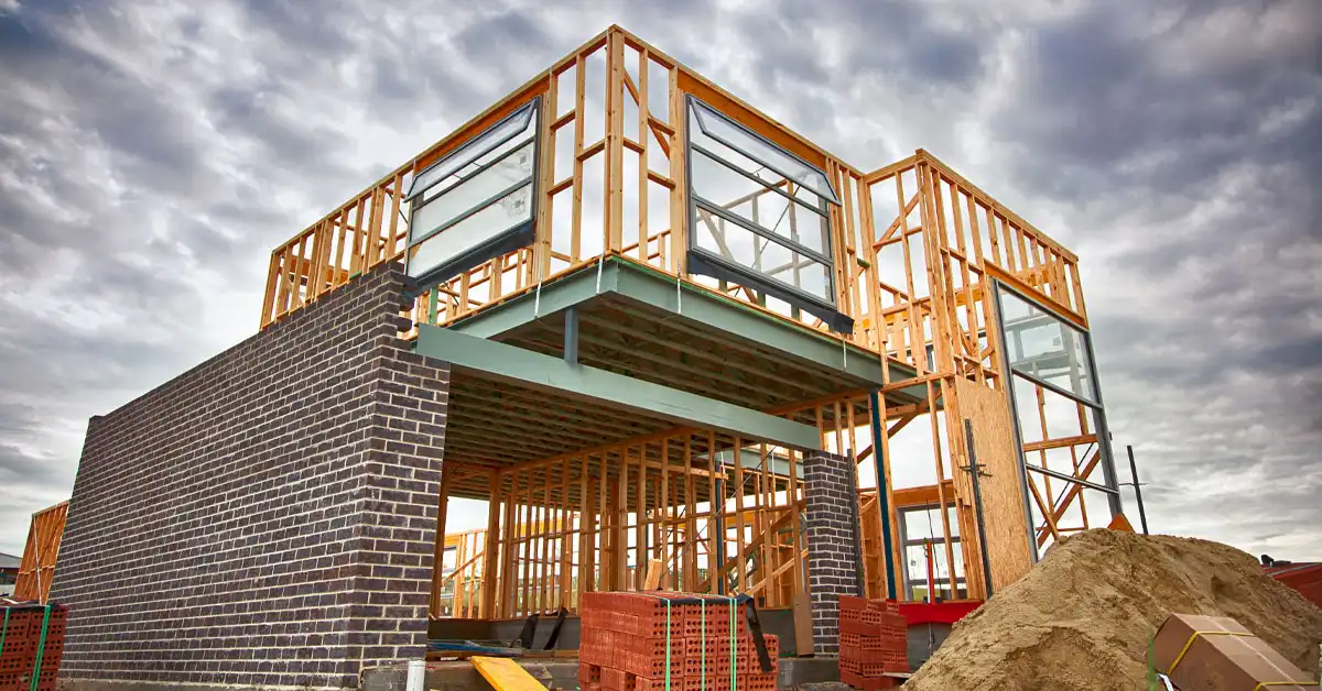 Partially constructed house with wooden frame and brick base under a cloudy sky. Piles of sand and bricks are visible, suggesting active construction.