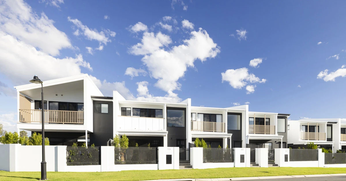 Modern row of townhouses under a bright blue sky with fluffy clouds. The white and black buildings feature balconies and well-kept greenery in front.