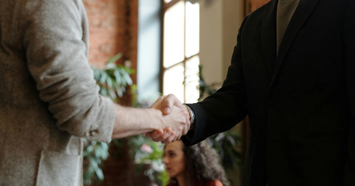 Two people shake hands in a warmly lit room with plants and a brick wall. A seated person with curly hair is blurred in the background, creating a professional atmosphere.