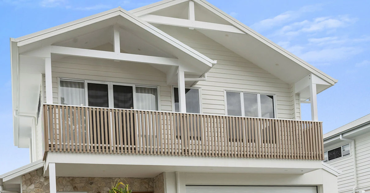 Modern two-story house with light beige wood siding, a balcony with vertical slats, and large windows under a clear blue sky. Cozy and inviting design.