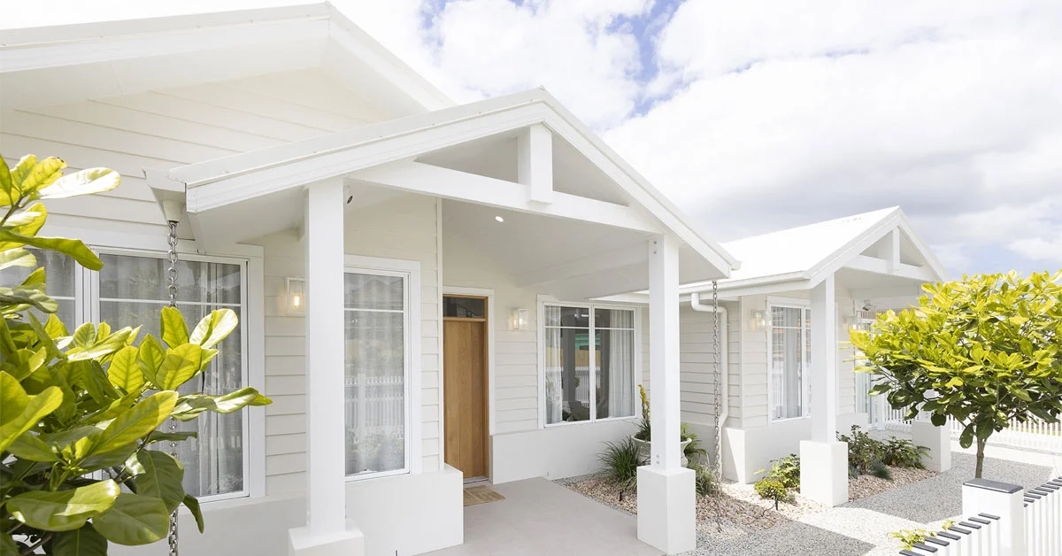 Modern, white single-story house with large windows and a wooden door. Surrounded by green plants and a white picket fence under a partly cloudy sky.